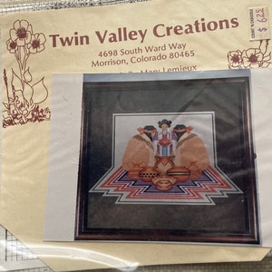 May include: A framed cross-stitch pattern featuring Native American figures and pottery. The artwork is in a dark wooden frame, with a geometric design at the base. The packaging includes the text "Twin Valley Creations".