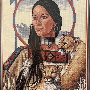Puede incluir: Obra de arte de punto de aguja enmarcada que representa a una mujer nativa americana con cabello largo y negro, vestida con atuendo tradicional. Se representan dos pumas junto a ella. La obra tiene un borde azul y rojo, y una cordillera es visible en el fondo.