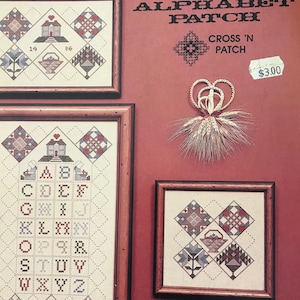 May include: Three framed cross-stitch samplers with a red background. The samplers feature geometric patterns, floral designs, and the alphabet. The text "ALPHABET PATCH" and "CROSS 'N PATCH" are visible. A decorative wheat and heart ornament is also present.