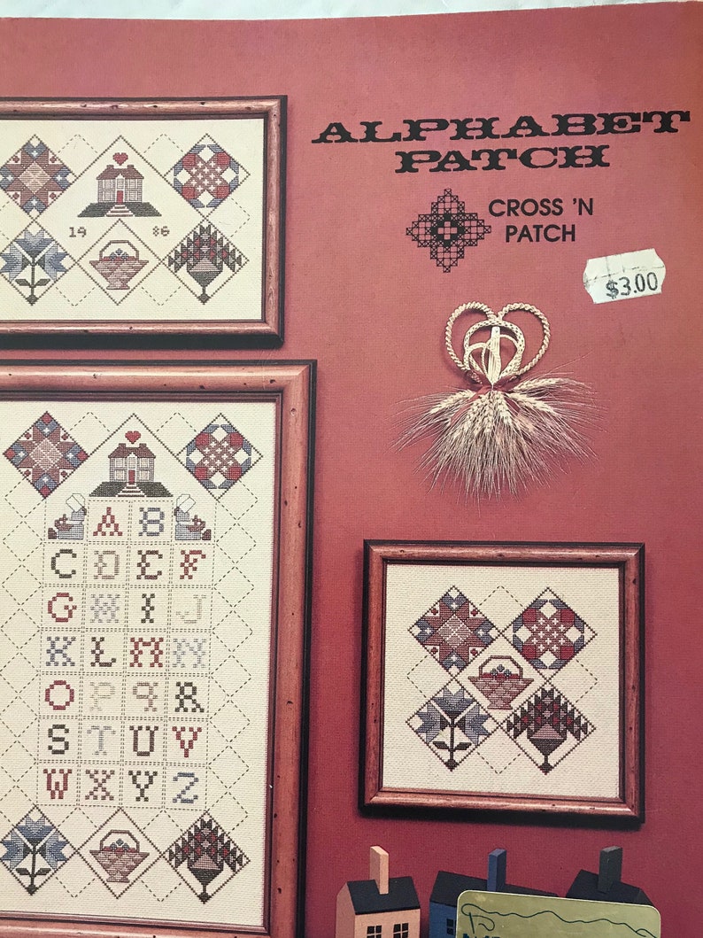 Op de afbeelding: Drie ingelijste kruissteekstalen tegen een rode achtergrond. De stalen tonen geometrische patronen, bloemmotieven en het alfabet. De tekst "ALPHABET PATCH" en "CROSS 'N PATCH" is zichtbaar. Een decoratief tarwe- en hartornament is ook aanwezig.