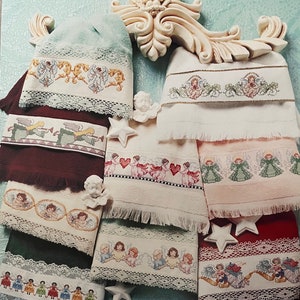 Puede incluir: Colecci&oacute;n de toallas bordadas con dise&ntilde;os de &aacute;ngeles. Las toallas presentan varios colores, incluyendo blanco, verde y burdeos, con patrones de punto de cruz detallados de &aacute;ngeles y adornos de encaje decorativos. La imagen tambi&eacute;n incluye figuritas de &aacute;ngeles decorativas y adornos en forma de estrella.