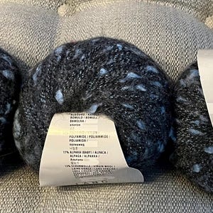 Puede incluir: Tres ovillos de hilo Lang Freya en una combinación de colores gris oscuro moteado y azul claro. Cada ovillo tiene una etiqueta con el nombre de la marca e información del producto. El hilo es de 50g y 205m.