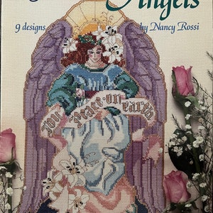 Op de afbeelding: Een kruissteekpatroonboek getiteld "Angels" van Nancy Rossi. De omslag toont een kruissteek engelontwerp met een vrouw in een blauwe jurk, omringd door paarse vleugels en witte lelies. De tekst "Your Peace on Earth" is zichtbaar.