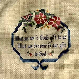 May include: Cross-stitch embroidery on cream fabric. The design features a blue, octagonal frame with floral accents in red, green, and yellow. Text within the frame reads, "What we are is God's gift to us. What we become is our gift to God."