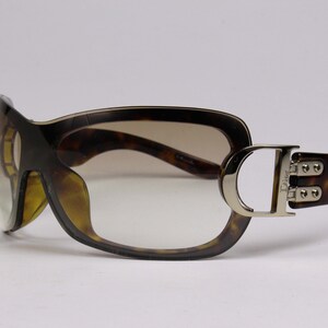May include: Brown and gold-toned sunglasses with a large, square frame and a tortoiseshell pattern. The sunglasses have a silver metal accent on the side with the word "Dior" engraved on it.