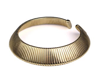 Puede incluir: Un collar dorado con un diseño acanalado. El collar de estilo gargantilla abierto tiene forma redondeada y está sobre un fondo blanco. El metal tiene un acabado brillante.