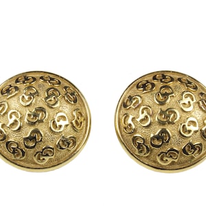 May include: A pair of gold-tone earrings with a circular design featuring a repeating pattern of the letter 'G'.