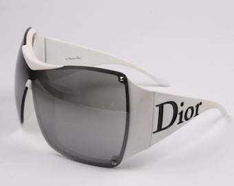 Peut inclure: Lunettes de soleil surdimensionnées avec une monture blanche et des verres teintés gris foncé. Les branches affichent le texte noir Dior, tandis que l'intérieur porte la mention Christian Dior. Des petits clous argentés ornent les coins.