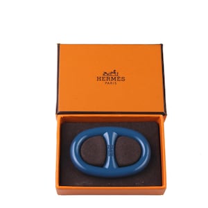 May include: A blue oval-shaped accessory rests inside an orange box. The box is open, revealing the product against a brown interior. The box has the brand name and logo printed on the lid.