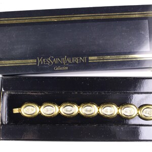 Puede incluir: Un brazalete dorado con piedras transparentes en una caja negra con el texto "Yves Saint Laurent Collection" en la tapa.