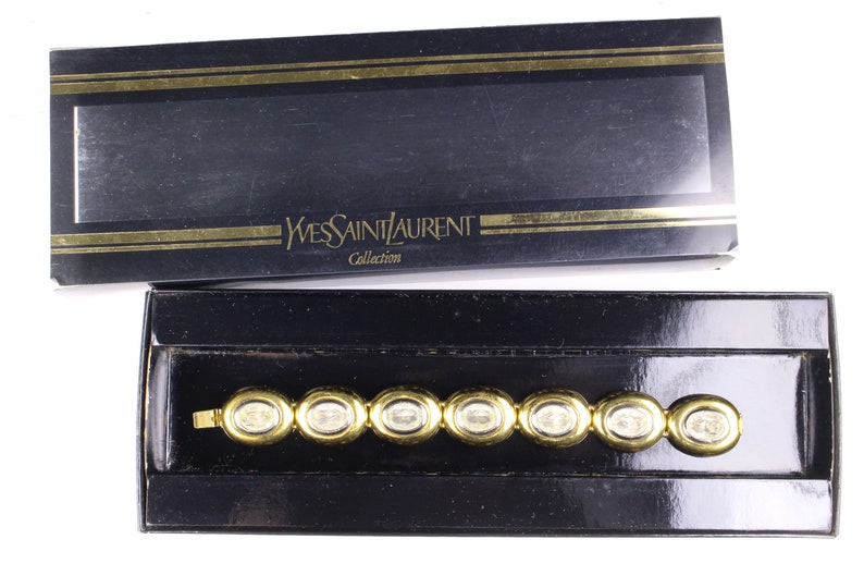 Puede incluir: Un brazalete dorado con piedras transparentes en una caja negra con el texto "Yves Saint Laurent Collection" en la tapa.