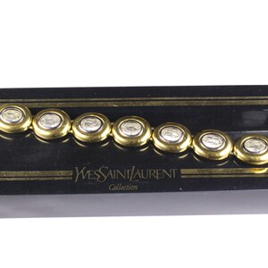 Puede incluir: Un brazalete dorado con piedras ovaladas transparentes. El brazalete est&aacute; en una caja negra con las palabras "Yves Saint Laurent Collection" impresas en la parte delantera.