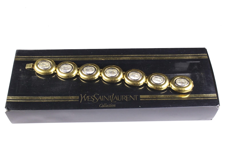 Puede incluir: Un brazalete dorado con piedras ovaladas transparentes. El brazalete est&aacute; en una caja negra con las palabras "Yves Saint Laurent Collection" impresas en la parte delantera.