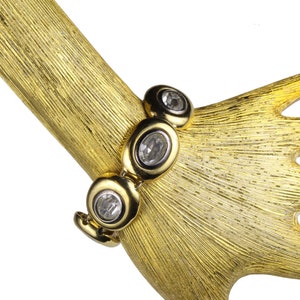 Puede incluir: Un brazalete dorado con piedras ovaladas transparentes. El brazalete est&aacute; en una mano de maniqu&iacute; dorada.