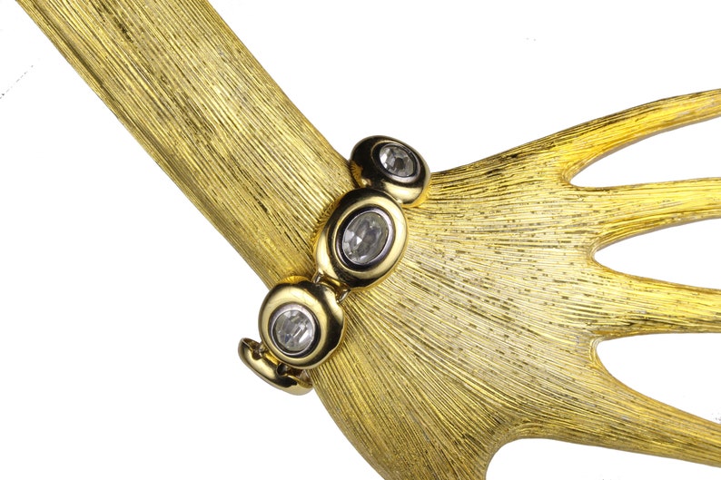 Puede incluir: Un brazalete dorado con piedras ovaladas transparentes. El brazalete est&aacute; en una mano de maniqu&iacute; dorada.