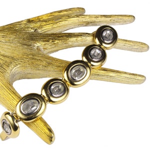 Puede incluir: Un brazalete de oro con piedras ovaladas transparentes engastadas en engastes de oro. El brazalete est&aacute; en una mano dorada.