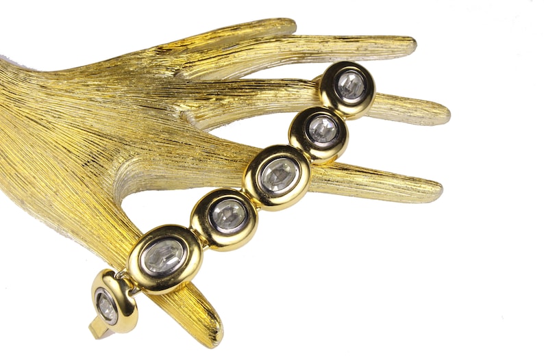 Puede incluir: Un brazalete de oro con piedras ovaladas transparentes engastadas en engastes de oro. El brazalete est&aacute; en una mano dorada.