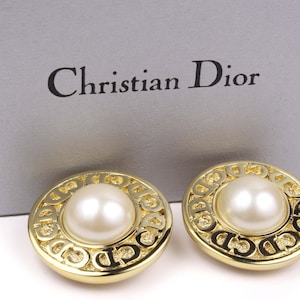 May include: Gold-tone clip-on earrings featuring a large faux pearl centre. The earrings have a circular design with the "CD" logo. The brand name "Christian Dior" is visible on the box in the background.