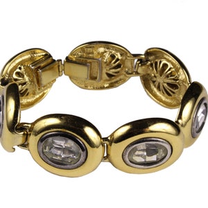 Puede incluir: Un brazalete de oro con piedras ovaladas transparentes engastadas en el centro de cada eslab&oacute;n. El brazalete tiene un cierre decorativo con un dise&ntilde;o floral.