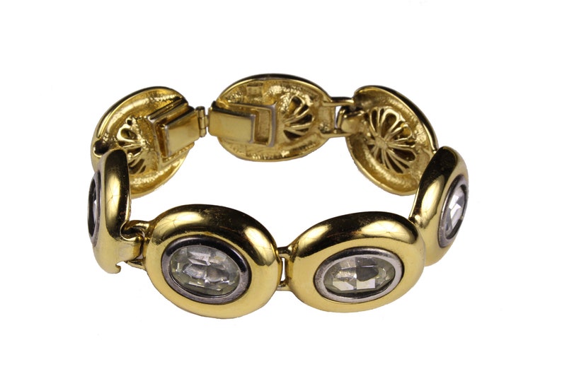 Puede incluir: Un brazalete de oro con piedras ovaladas transparentes engastadas en el centro de cada eslab&oacute;n. El brazalete tiene un cierre decorativo con un dise&ntilde;o floral.