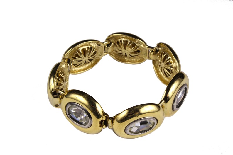 Puede incluir: Un brazalete dorado con eslabones de forma ovalada, cada uno con un brillante de cristal transparente. Los eslabones est&aacute;n conectados por un mecanismo de bisagra.