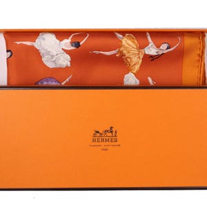 May include: An orange box with a black trim contains a silk scarf. The scarf features a ballet dancer design in white, gold, and purple. The box has the Hermes logo and text at the bottom.