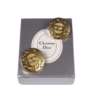 May include: A pair of gold-coloured Christian Dior earrings. The earrings are round with a textured border and the CD logo in the centre. They are displayed on a silver box with the Christian Dior logo.
