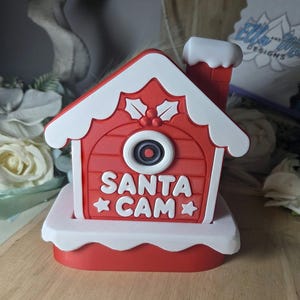 Könnte beinhalten: Ein rot-weißes, hausförmiges Santa Cam Ornament mit Schornstein, einer zentralen Kameralinsengrafik und dem Text SANTA CAM mit zwei Sternen. Dach und Sockel haben weiße, schneeartige Details, und oben befinden sich Stechpalmenblätter mit Beeren.