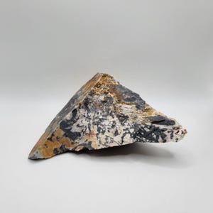 May include: A triangular-shaped, raw stone specimen with a mix of colors. The stone features black, white, tan, and brown hues, creating a marbled effect. The rough texture and natural form suggest a geological or mineral specimen.