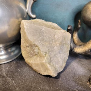 May include: A rough-hewn, light gray stone with a textured surface. The stone has subtle green and tan hues and is positioned near metallic decorative objects. The stone's irregular shape and color variations suggest a natural origin.