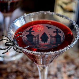 May include: Two martini glasses filled with a red liquid, rimmed with sugar, and decorated with a spider. The liquid in the glasses features a Halloween-themed image of a graveyard with a red moon.