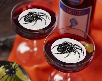 Puede incluir: Dos copas de vino tinto con una pegatina redonda blanca en cada copa que representa una araña negra. Las copas están sobre un mantel rojo y naranja con una araña y un murciélago en la mesa. El vino es de la marca Barefoot.