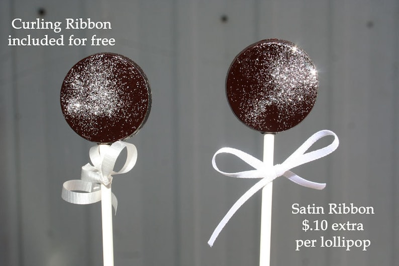 Puede incluir: Dos piruletas de chocolate con palos blancos y lazos de cinta blanca. La piruleta de la izquierda incluye un lazo de cinta rizada gratis. La piruleta de la derecha tiene un lazo de cinta de sat&eacute;n por 1,10 &euro; extra por piruleta.