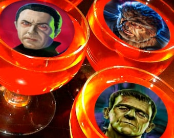 Puede incluir: Cuatro vasos de chupito de vidrio transparente llenos de gelatina naranja. Cada vaso tiene una pegatina redonda en el fondo con una imagen diferente en blanco y negro de un monstruo clásico de película de terror. Los monstruos son Drácula, el Hombre Lobo, el Monstruo de Frankenstein y la Criatura de la Laguna Negra.