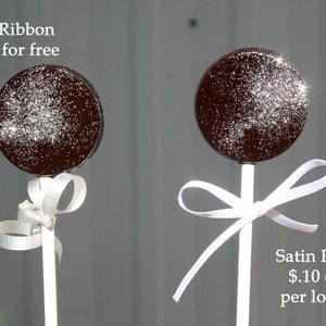 May include: Two chocolate lollipops with white sprinkles on top. The lollipop on the left has a white curling ribbon tied around the stick. The lollipop on the right has a white satin ribbon tied around the stick. The text "Curling Ribbon included for free" is at the top left of the image. The text "Satin Ribbon $1.10 extra per lollipop" is at the bottom right of the image.