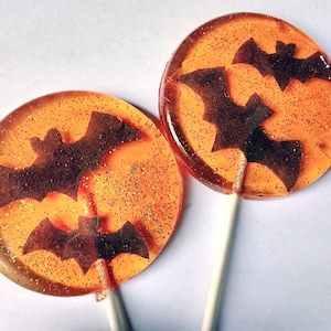 May include: Two orange and brown lollipops with black bat silhouettes. The lollipops have a sparkly, glittery texture.