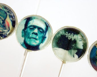 May include: Four round lollipops with images of classic horror movie characters. The lollipops are on white sticks and have a clear, glossy finish. The images are of Frankenstein's monster, the Creature from the Black Lagoon, Dracula, and the Wolfman.