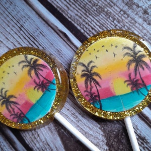 May include: Two round lollipops with gold glitter edges and a sunset scene with palm trees and birds. The lollipops are on a wooden surface.