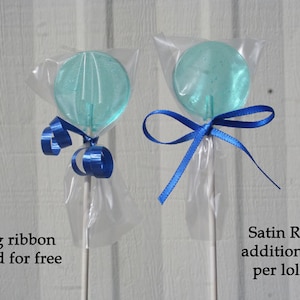 May include: Two clear, round, blue lollipops on white sticks, wrapped in cellophane. One is tied with blue curling ribbon, the other with a blue satin ribbon. The text reads "Curling ribbon included for free" and "Satin Ribbon additional $.10 per lollipop."