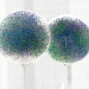 May include: Two round lollipops with green, blue, and purple glitter embedded in clear candy. The lollipops are on white sticks.