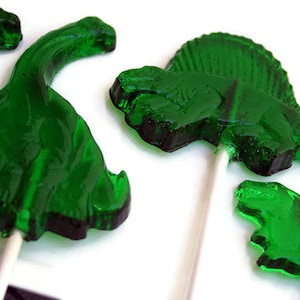 May include: Four green dinosaur lollipops on sticks. Two of the lollipops are shaped like a dinosaur with a spiky back, one is shaped like a long-necked dinosaur, and one is shaped like a dinosaur with a short neck and a long tail.