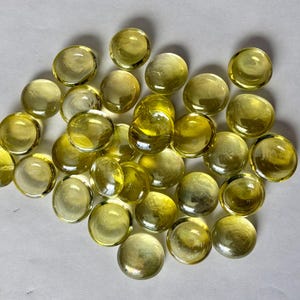 May include: A collection of approximately 30 yellow-green glass marbles. The marbles are round and vary slightly in size. They have a translucent quality, with some appearing more opaque than others. The marbles are scattered on a white surface.