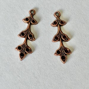 May include: Two identical copper-toned metal charms shaped like elongated, stylized leaves or branches. Each piece features a vertical stem with three tiers of symmetrical, rounded leaf-like segments, each containing a small circular cutout.