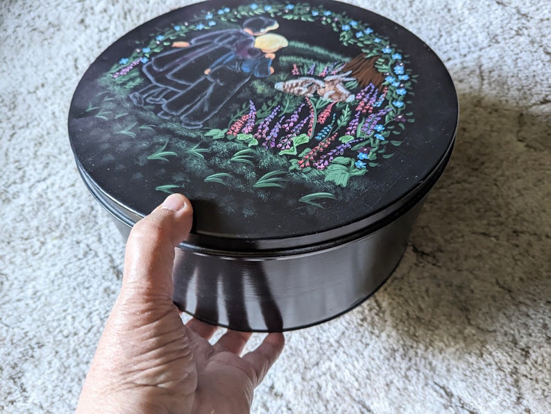 May include: A black round tin with a painted scene of two people in a field of flowers. The lid of the tin has a floral design with a wreath of flowers around the edge.