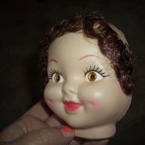 May include: A doll head with brown hair, painted on eyelashes, and yellow eyes. The doll head is made of plastic and has a painted-on smile.