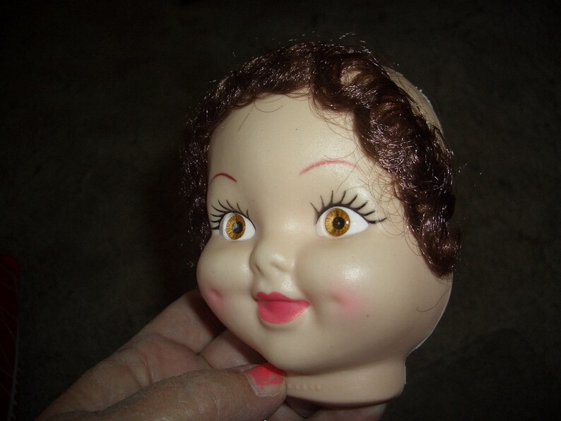 May include: A doll head with brown hair, painted on eyelashes, and yellow eyes. The doll head is made of plastic and has a painted-on smile.