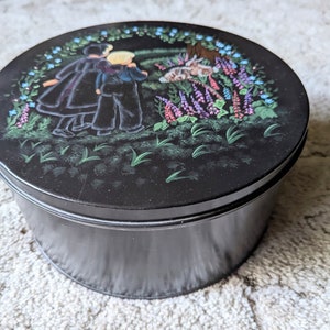 May include: Black round tin with a painted scene of three people walking in a field with flowers. The people are wearing black clothing and the flowers are pink, purple, and blue.