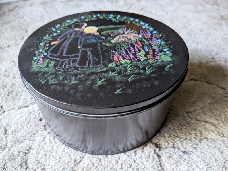 May include: Black round tin with a painted scene of three people walking in a field with flowers. The people are wearing black clothing and the flowers are pink, purple, and blue.