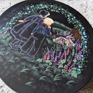 May include: A round wooden plate with a black background and a painted scene of two figures in black clothing walking through a field of purple and blue flowers. Two rabbits are in the foreground.