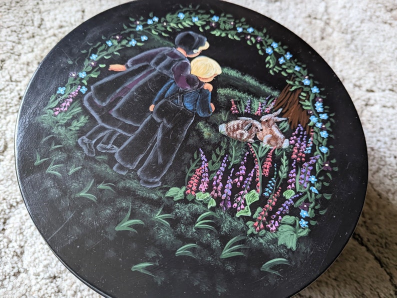 May include: A round wooden plate with a black background and a painted scene of two figures in black clothing walking through a field of purple and blue flowers. Two rabbits are in the foreground.
